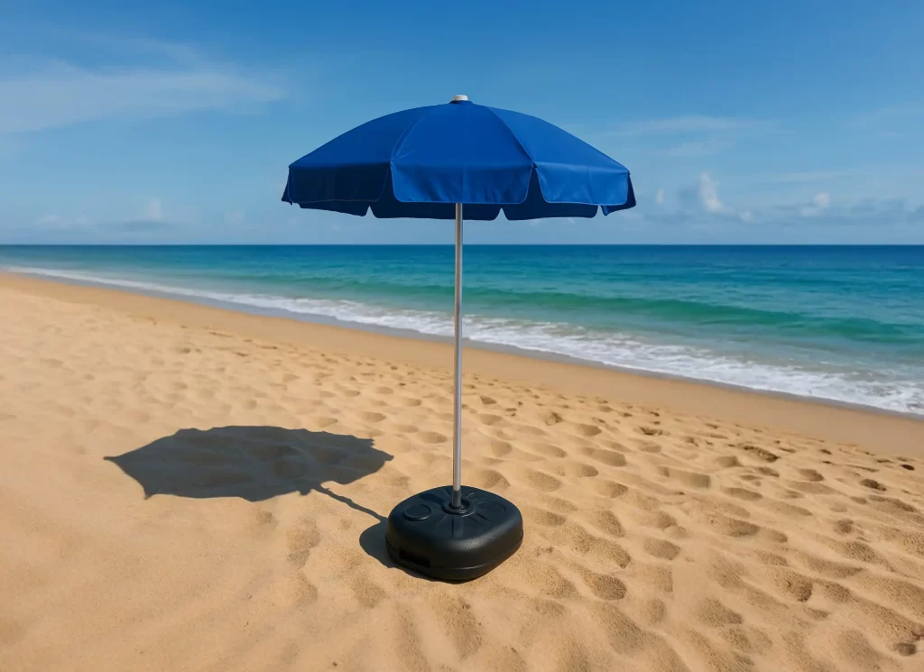 Guarda-sol azul montado em uma base plástica preta sobre a areia da praia em um dia ensolarado, com o mar e o céu azul ao fundo.